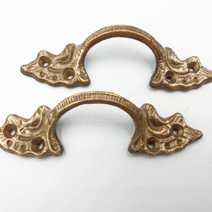 Könnte beinhalten: Zwei antike Schubladengriffe in Bronzefarbe. Jeder Griff hat einen gebogenen Griff und dekorative Endstücke mit drei Befestigungslöchern. Die Griffe sind aus Metall und haben eine Vintage-Ästhetik.