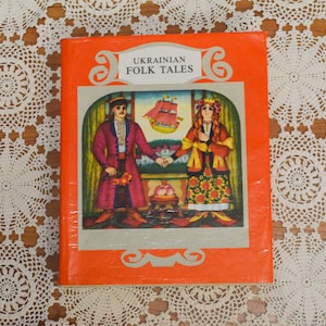 Pode incluir: Um livro vintage intitulado "Ukrainian Folk Tales" com uma capa laranja. A capa apresenta uma ilustração colorida de um homem e uma mulher em trajes tradicionais, de mãos dadas. Um navio navega ao fundo. O livro está sobre uma toalha de renda branca.