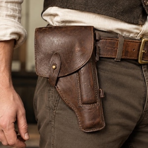 Puede incluir: Una funda de cuero marrón oscuro con cierre de solapa y un botón de latón, sujeta a un cinturón ancho de cuero marrón. La funda presenta un diseño estructurado con un bolsillo lateral, asegurada a la cintura sobre pantalones de color oliva.