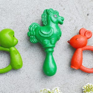 May include: Three colorful baby rattles in the image. A green monkey rattle, a green horse rattle, and an orange mouse rattle. The rattles are made of plastic and have a ring handle.