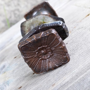 May include: A stack of three square-shaped cabinet knobs with a dark, metallic bronze finish. Each knob features a central floral relief pattern surrounded by a textured grid design. The knobs are arranged vertically, showing the top face clearly.