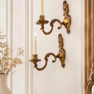 May include: Two ornate gold-toned wall sconces mounted vertically, each featuring a curved arm and a decorative backplate. Each sconce holds a single slender white taper candle with a lit flame, creating a warm illumination against the wall.