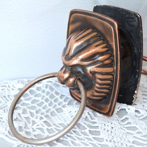 Pode incluir: Um aldravão de porta em metal com tom de cobre, apresentando um design de cabeça de leão esculpida com um anel circular proeminente na boca. A placa traseira retangular tem um acabamento escuro e desgastado com um logótipo circular em relevo.