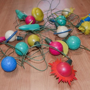 May include: A collection of vintage Christmas lights with various shapes and colors. The lights include spheres, rockets, and sunbursts in shades of red, yellow, blue, and green. The lights are strung together with green wire and are scattered on a wooden surface.