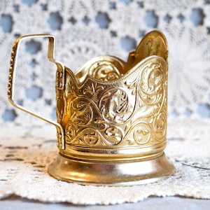 Può includere: Porta tazza dorato con manico e motivi floreali e a volute. Il supporto ha una base rotonda e il manico è semplice e curvo. Lo sfondo è una tovaglietta di pizzo bianco.