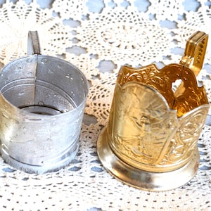 Può includere: Due portabicchieri vintage in metallo. Uno è argentato con un manico e perforazioni decorative. L'altro è dorato con dettagli floreali ornati e un manico. Entrambi sono progettati per contenere una tazza o un bicchiere.