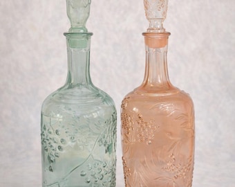 May include: Two decorative glass decanters. One is a pale green color, and the other is a soft peach. Both have a grape vine design and a decorative stopper. These decanters are ideal for serving wine or other beverages.