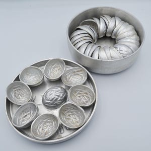 May include: A set of vintage aluminum molds for making small cakes or pastries. The set includes a round container filled with molds, and a tray with several molds arranged around a central, larger mold. The molds have a decorative, textured design.