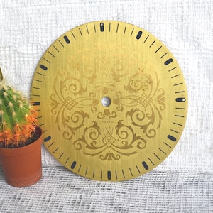 May include: A circular gold-toned clock face featuring an intricate etched scrollwork pattern and black hour markers along the perimeter. The center includes a small circular hole and the Cyrillic text "КВАРЦ" printed in black ink.