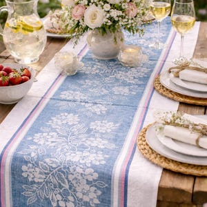 May include: A table setting with a blue floral table runner, white plates, and wine glasses. A bouquet of white and pink flowers sits in a white vase. Strawberries in a white bowl and a pitcher of water with lemon slices are also on the table.