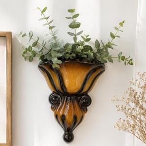 May include: A wall-mounted ceramic planter featuring a glossy finish with a gradient of deep amber and dark brown tones. The vessel has a flared top edge and a decorative, sculpted base with scroll details, containing a green eucalyptus plant.