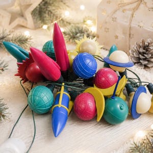 May include: A string of decorative holiday lights featuring various space-themed shapes including rockets, planets with rings, moons, and globes in vibrant colors like red, blue, yellow, and teal, connected by a thin dark green electrical wire.