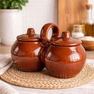 May include: Two brown ceramic cooking pots with lids and a handle. The pots have a glossy finish and sit on a woven placemat. The background includes a wooden chopping board and a white vase with greenery.