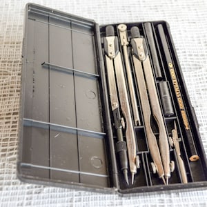 May include: An open black case containing drafting tools, including two compasses, a divider, and a pencil. The tools are silver-colored and appear to be vintage. The case is rectangular and has a ridged interior.