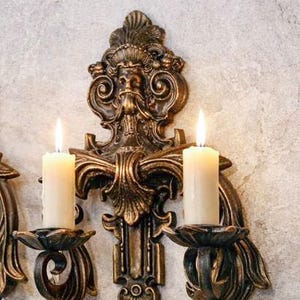 Puede incluir: Aplique de pared ornamentado, color bronce, con dos velas encendidas. El aplique presenta un diseño esculpido detallado con una cara central y adornos. Las velas son blancas y cilíndricas, proporcionando un brillo cálido.