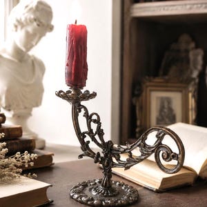 May include: A dark bronze-toned metal candle holder with an ornate, curved, and scrollwork design. The base is circular with a textured edge. A single, thick, deep red pillar candle with visible wax drips sits in the holder, topped with a flame.