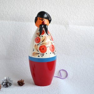 May include: A traditional wooden toy Cossack figure. It features a red base, a white body adorned with floral patterns, and an orange head with a black hat and mustache. A purple string is attached.