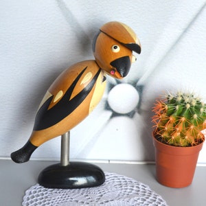 Puede incluir: Figura de pájaro de madera con diseño negro y dorado, montada sobre una varilla metálica y base negra. El pájaro tiene pico, ojos y una pequeña lengua rosa. Un pequeño cactus en maceta con espinas naranjas está cerca.