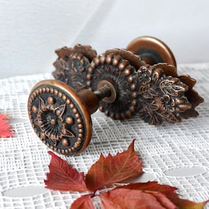 May include: A copper-toned metal drawer knob featuring a circular face with a central floral motif surrounded by a beaded border. The base plate is elongated with an intricate leaf and flower pattern, and a threaded metal bolt extends from the back.