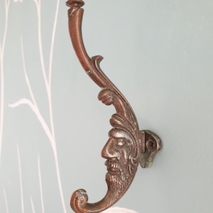 May include: A metallic, copper-toned wall hook featuring a sculpted human face with a beard integrated into the design. The hook has an elongated, curved neck that extends upwards to a rounded knob, with a smaller hook at the base for hanging items.