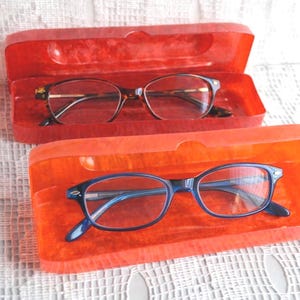May include: Two pairs of eyeglasses in open red cases. One pair has a brown tortoiseshell frame, and the other has a blue frame. The cases are rectangular and lined with a soft, orange material. The glasses are on a white, textured surface.