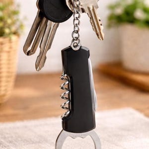 Puede incluir: Un llavero multiherramienta negro y plateado con un abridor de botellas, un sacacorchos y otras herramientas. Varias llaves cuelgan del llavero. El llavero está sobre una superficie clara.