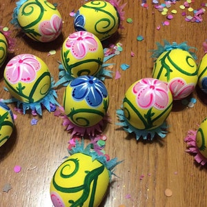 May include: A collection of yellow Easter eggs decorated with green vines, pink and blue flowers. The eggs are sitting on a wooden surface with colourful confetti.