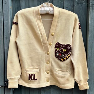 May include: A cream-colored knit cardigan featuring a V-neckline, long sleeves with ribbed cuffs, and a button-down front. The garment is decorated with a large dark brown bulldog patch on the left chest, the letters KL on the right pocket, and patches.