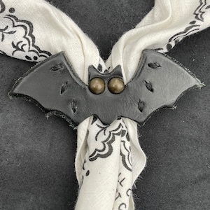 May include: A black leather bat-shaped scarf holder with two brass-colored buttons. The holder is attached to a white bandana with a black floral pattern.