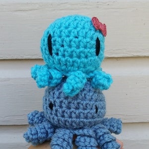 May include: Two crocheted octopus amigurumi toys, one blue and one light blue, with black eyes and a pink bow on the top of the blue octopus. The text "Pi & Pippi the Octopi" is above the toys and "CROCHET PATTERN by Eyeloveknots" is below the toys.