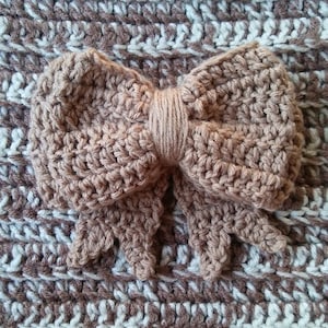 Könnte beinhalten: Eine gehäkelte Schleife in Hellbraun mit strukturierter Optik liegt auf einem Hintergrund aus braun-weißem Häkelwerk. Die Schleife hat ein doppellagiges Design und ist mittig platziert. Der Text unten lautet "Double Layered Bow CROCHET PATTERN".