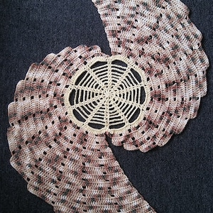 May include: A crocheted table runner in shades of brown, beige, and white. The runner is made up of two half-circles joined together, with a circular design in the center. The text "Ila's Table Runner" is at the top of the image, and the text "CROCHET PATTERN by EyeLoveKnots" is at the bottom.