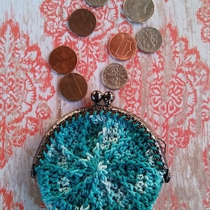 Può includere: Un portamonete all'uncinetto turchese e bianco con chiusura in metallo, circondato da monete. La borsa ha una forma rotonda e un motivo a vortice strutturato. Lo sfondo presenta un motivo floreale e il testo "Whirlpool Coin Purse CROCHET PATTERN By EyeLoveKnots".