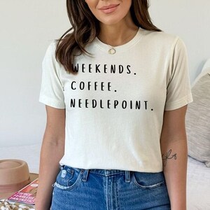 May include: A woman wearing a light beige t-shirt with the text "Weekends. Coffee. Needlepoint." printed in black. She is wearing blue jeans and has a small tattoo on her wrist.