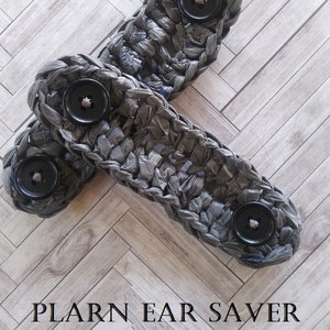 May include: Two gray crocheted ear savers with black buttons. The ear savers are made with plastic yarn and are laid out on a woodgrain surface. The text "PLARN EAR SAVER CROCHET PATTERN" is at the bottom of the image.
