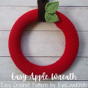 Può includere: Una ghirlanda di mele di uncinetto rossa con foglie di uncinetto verdi. La ghirlanda è appesa su uno sfondo di legno bianco. Ghirlanda di mele facile. Modello di uncinetto facile di EyeLoveKnots.