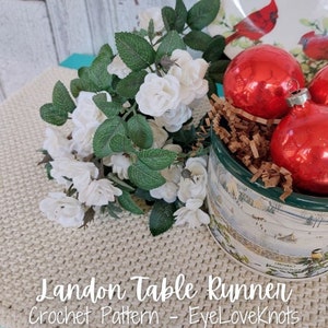 May include: A crocheted table runner in a beige color with the text "Landon Table Runner Crochet Pattern - EyeLoveKnots" on the bottom. A bouquet of white roses and a tin can filled with red Christmas ornaments are on top of the runner.