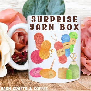 May include: White ceramic mug with a brown "Surprise Yarn Box" text and colorful illustrations of yarn balls, needles, scissors, and wool.