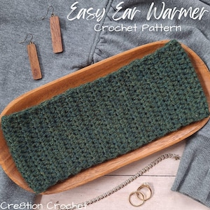 May include: A green crocheted ear warmer sits in a wooden tray, with the text "Easy Ear Warmer Crochet Pattern". Wooden earrings, a silver bracelet, and gold rings are also in the image.