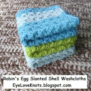 Op de afbeelding: Een stapel van vier gehaakte washandjes in blauw, groen en wit. De washandjes zijn gemaakt met een schuine schelpensteek. De tekst "Robin's Egg Slanted Shell Washcloths EyeLoveKnots.blogspot.com" is zichtbaar onderaan de afbeelding.