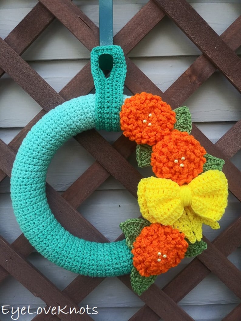 Puede incluir: Una corona de crochet de color turquesa con un lazo amarillo y flores de crochet de color naranja. La corona est&aacute; colgada en una celos&iacute;a de madera.