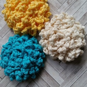 May include: Three handmade crochet facial scrubbies in yellow, white, and turquoise blue. The scrubbies are round and textured, with the text "ENDLESS SUMMER FACIAL SCRUBBIES CROCHET PATTERN" at the bottom. The background is a light wood pattern.