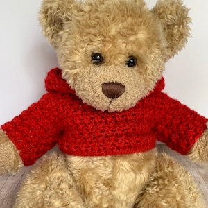 May include: A light brown teddy bear wearing a red crocheted hoodie. The bear is sitting on a wooden surface.
