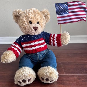 May include: A plush teddy bear wearing a patriotic sweater with red, white, and blue stripes and a pair of blue denim jeans. The bear is holding a small American flag, celebrating a national holiday.