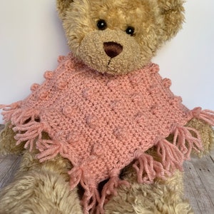 May include: A pink crocheted poncho with a textured bobble stitch and fringe trim. The poncho is worn by a brown teddy bear.