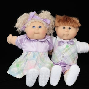 May include: Two vintage Cabbage Patch Kids dolls. The girl doll has blonde curly hair and wears a lavender dress with a plaid skirt. The boy doll has brown hair and wears a plaid vest and shorts. Both dolls have blue eyes.