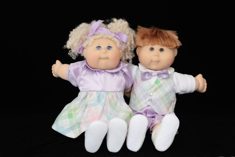 May include: Two vintage Cabbage Patch Kids dolls. The girl doll has blonde curly hair and wears a lavender dress with a plaid skirt. The boy doll has brown hair and wears a plaid vest and shorts. Both dolls have blue eyes.