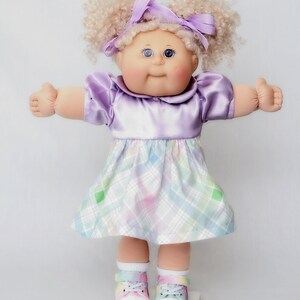 May include: A vintage doll with blonde curly hair styled in pigtails, adorned with a purple bow. The doll wears a lavender satin dress and a plaid skirt in pastel colors. White shoes with rainbow accents complete the look.