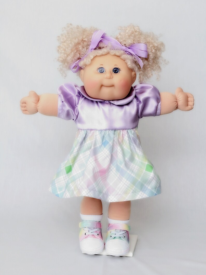 May include: A vintage doll with blonde curly hair styled in pigtails, adorned with a purple bow. The doll wears a lavender satin dress and a plaid skirt in pastel colors. White shoes with rainbow accents complete the look.
