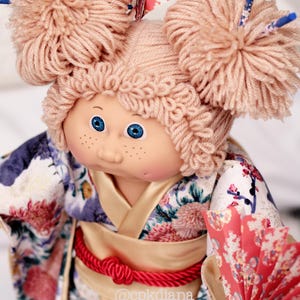 Op de afbeelding: Pop met blauwe ogen en sproeten, gekleed in een bloemenkimono met een rode touwriem. De pop heeft garen haar gestyled in twee grote knotjes en houdt een rode papieren waaier vast. Het haar van de pop is versierd met decoratieve stokjes.